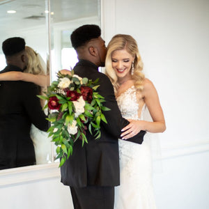 A bride and groom embrace while the bride holds a cascading bridal bouquet with burgundy and white flowers