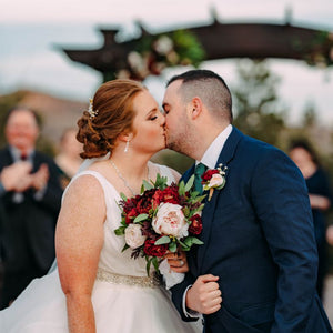 A bride and groom kiss in front of Willow Ridge Manor's arbor, the bride is holding her burgundy and blush bridal bouquet 