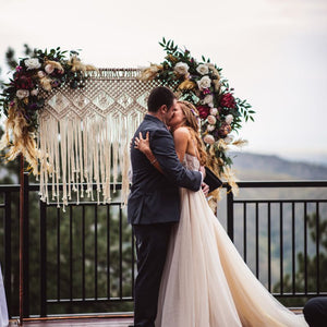 A couple shares their first kiss as husband and wife in front of a rectangular copper arch with macrame and two arch swags with boho inspired flowers and lots of feathers! 