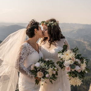 Two brides kiss on the top of a mountain holding bouquets made of cream and blush flowers with greenery. One of the bouquets is unstructured and the other is a cascading bouquet.