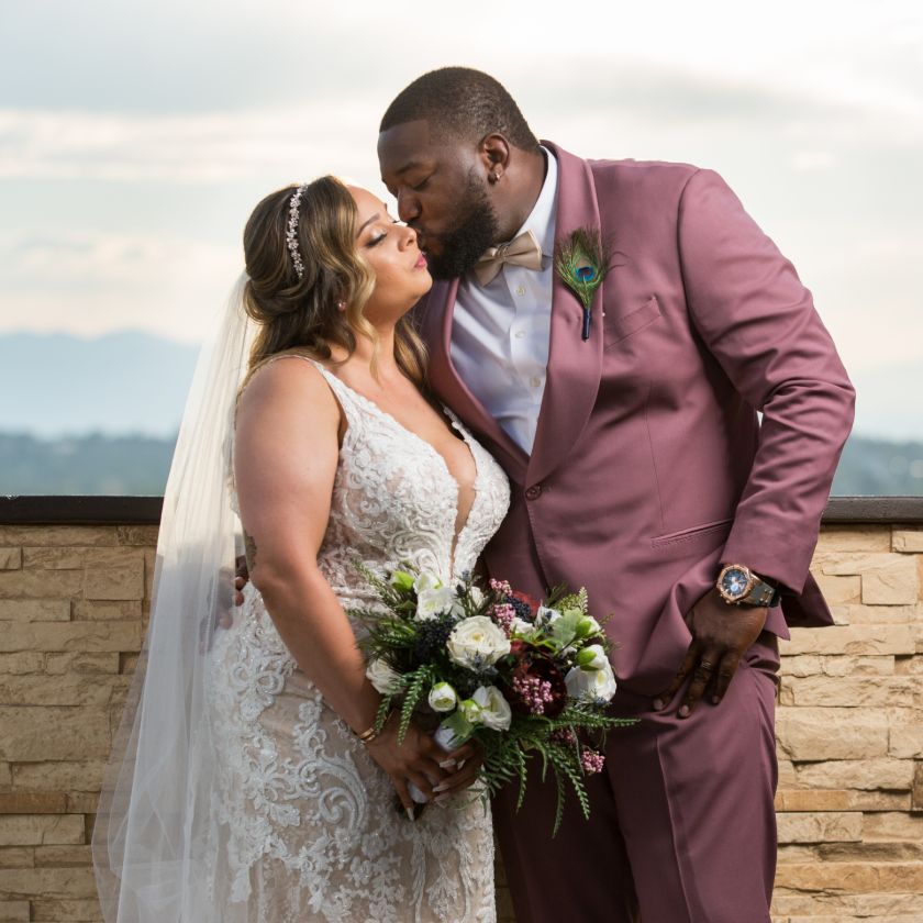 A groom kisses his bride while wearing a dusty rose suit, and his bride is holding a matching bridal bouquet of roses, ranunculus, and greenery. 