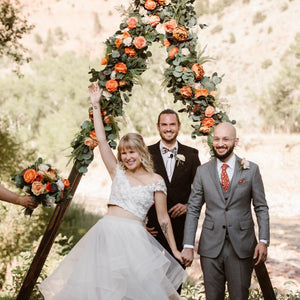 A couple who has just gotten married stands in front of their wooden triangle arch adorned with orange roses, peonies, lotus pods, and greenery. Compass Rose Floral can create the perfect arch flowers for the wedding of your dreams! 